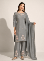 A woman models a three-piece gray ethnic suit set featuring a kurta with floral embroidery, matching wide-leg trousers, and a dupatta, set against a plain light beige studio background.
