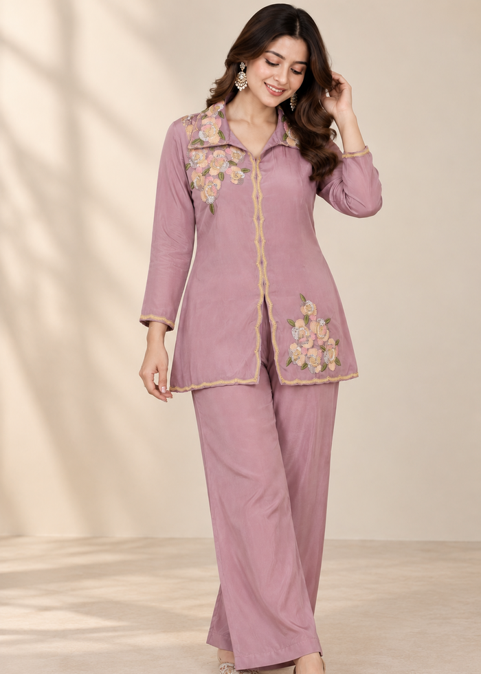 A woman modeling a dusty pink two-piece kurta set with floral embroidery and gold trim, standing against a neutral beige background.