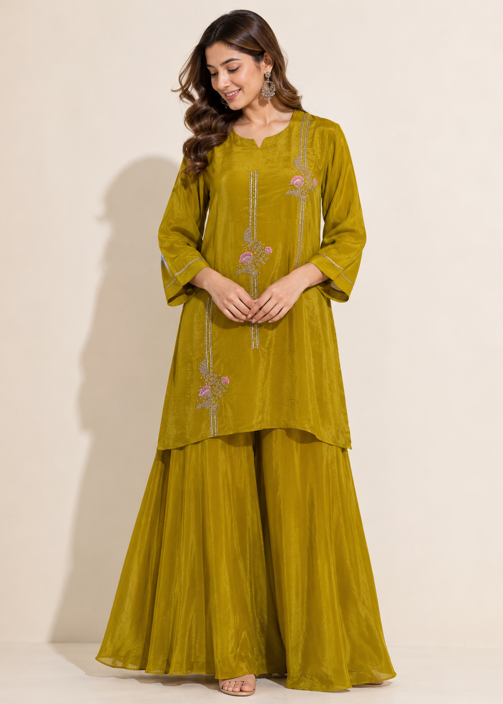 A woman modeling a three-piece olive green traditional outfit with floral embroidery on the kurta and matching wide-leg pants against a plain beige studio background.