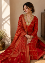 A woman wearing a red traditional outfit with gold embroidery and a matching dupatta, kneeling on a beige floor in a warm, sunlit room with a wooden cabinet in the background.