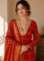 A woman wearing a red and gold traditional Indian outfit with intricate gold embroidery around the neckline, standing in a warm-toned indoor setting.