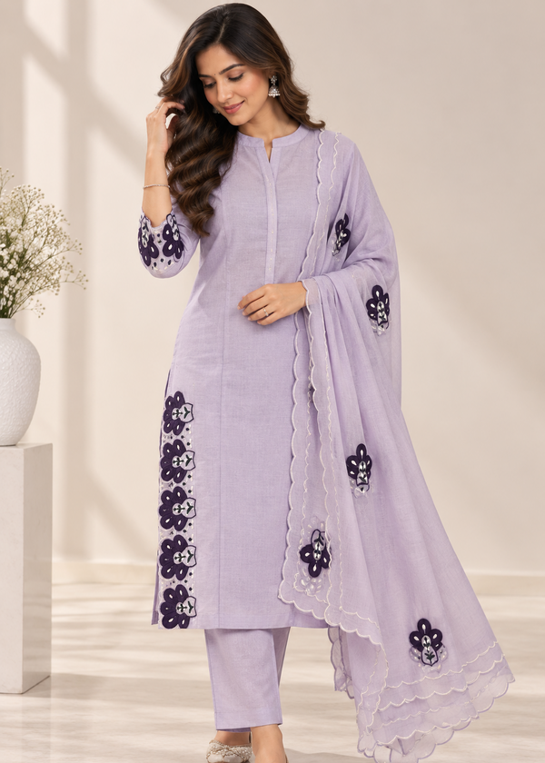 A woman models a light lavender purple ethnic salwar kameez suit with dark purple floral embroidery and a matching dupatta, set against a warm beige background.