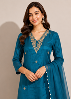 A woman modeling a teal blue traditional Indian salwar kameez suit with gold embroidery on the neckline and scattered motifs on the fabric, paired with a matching sheer dupatta.