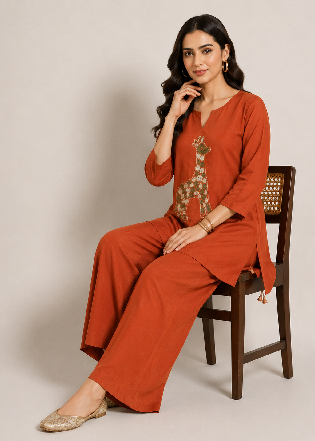 A woman sits on a wooden chair wearing a rust-orange two-piece kurta set with a giraffe embroidery detail on the chest. The set includes a long-sleeve V-neck top and matching wide-leg trousers, styled with gold flat shoes against a neutral beige background.