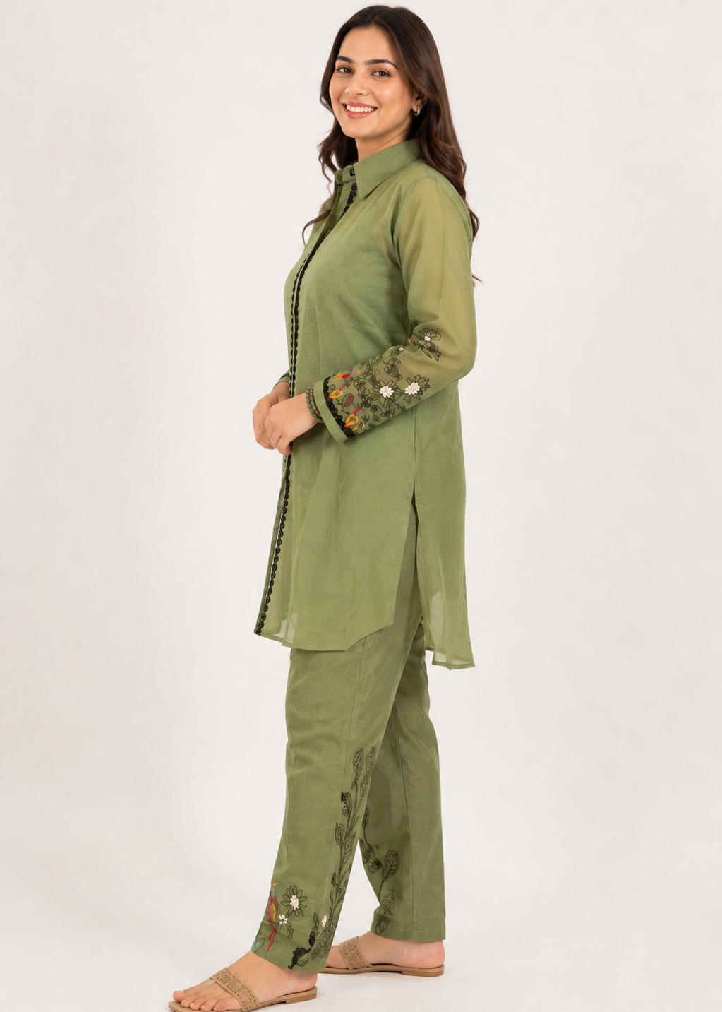 A woman modeling a sage green two-piece kurta set with floral embroidery on the sleeves and hem, standing against a plain white studio background.