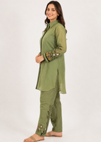 A woman modeling a sage green two-piece kurta set with floral embroidery on the sleeves and hem, standing against a plain white studio background.