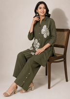 A woman wearing an olive green kurta set with white floral embroidery sits on a wooden chair against a neutral beige background.