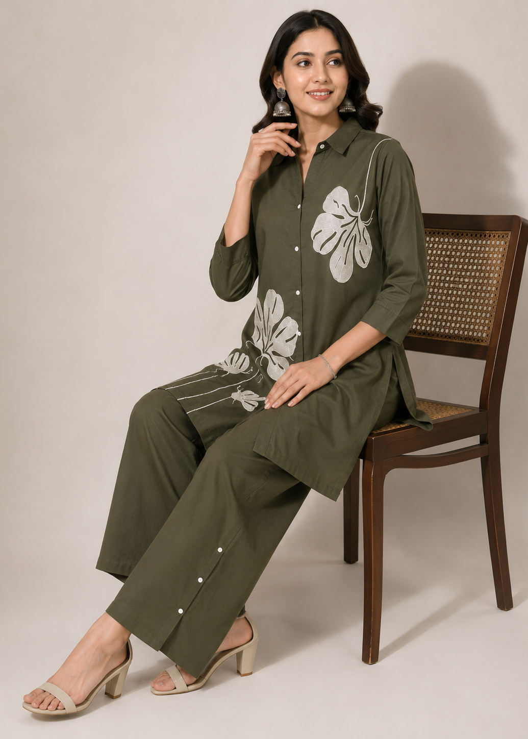 A woman wearing an olive green kurta set with white floral embroidery sits on a wooden chair against a neutral beige background.