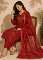 A woman seated on a woven chair wearing a deep red embroidered salwar kameez suit with a matching sheer dupatta. The outfit features gold and green floral embroidery, and the scene is styled with a metallic vase of dried flowers against a beige wall.