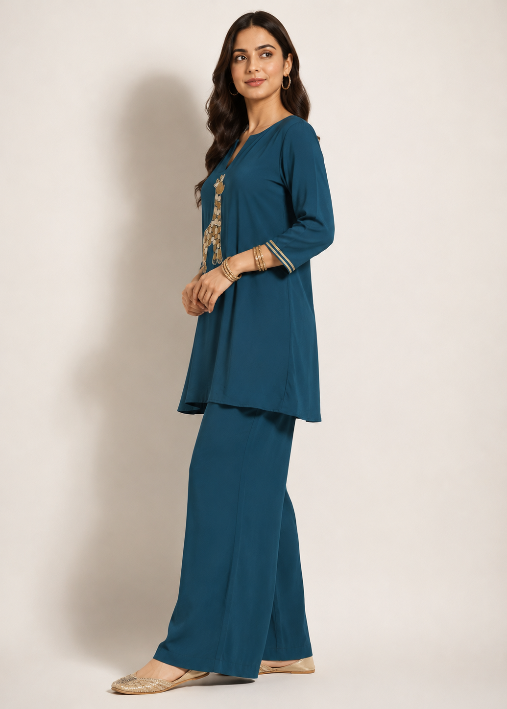 A woman modeling a teal blue kurta and wide-leg pant set with gold embroidery on the chest, standing against a plain beige studio background.