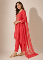 A woman models a coral pink three-piece traditional Indian outfit consisting of a kurta with gold embroidery, matching trousers, and a sheer matching dupatta, set against a plain beige studio background.