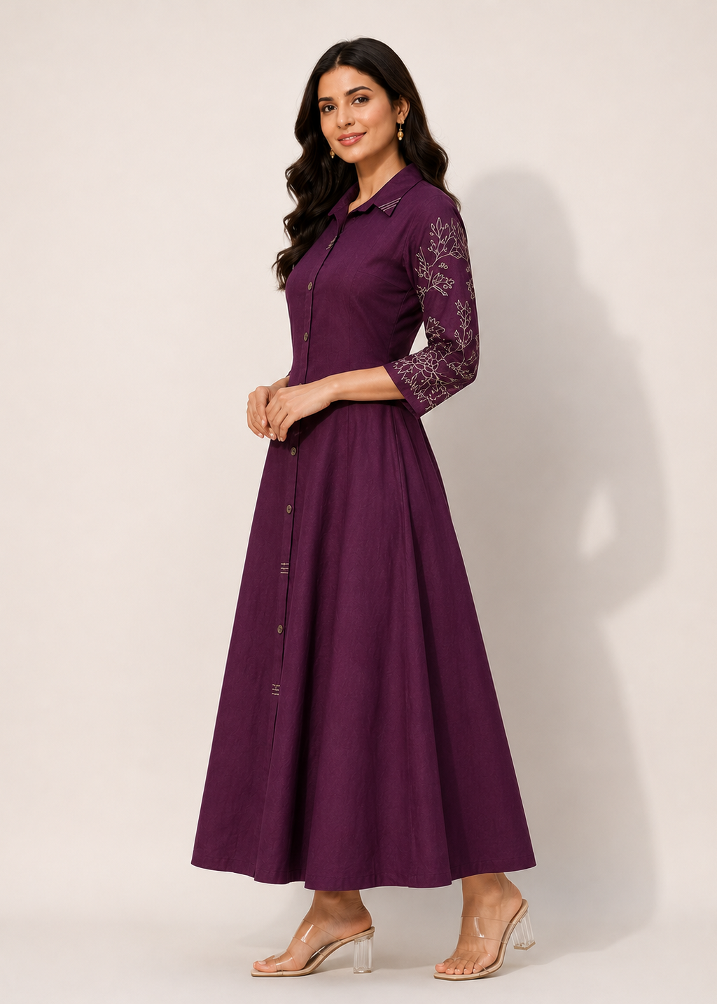 A woman modeling a long, sleeveless purple maxi dress with three-quarter sleeves featuring purple floral lace embroidery, a button-down front, and a flared skirt, set against a plain light beige studio background.