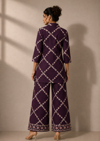A woman shown from the back wearing a deep purple two-piece outfit with a white geometric lattice pattern and matching border detailing on the pants, set against a warm beige wall with soft window shadows.
