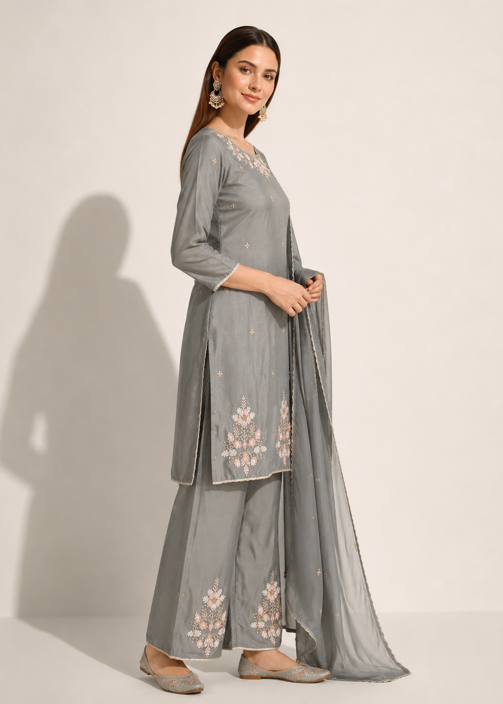 A woman modeling a light gray three-piece traditional outfit with floral embroidery, consisting of a kurta, wide-leg pants, and a matching dupatta, set against a plain off-white background.