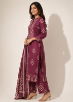 A woman modeling a three-piece traditional Indian outfit in deep plum purple with off-white geometric and chevron patterns, featuring a long kurta, wide-leg pants, and a matching dupatta, set against a plain beige studio background.