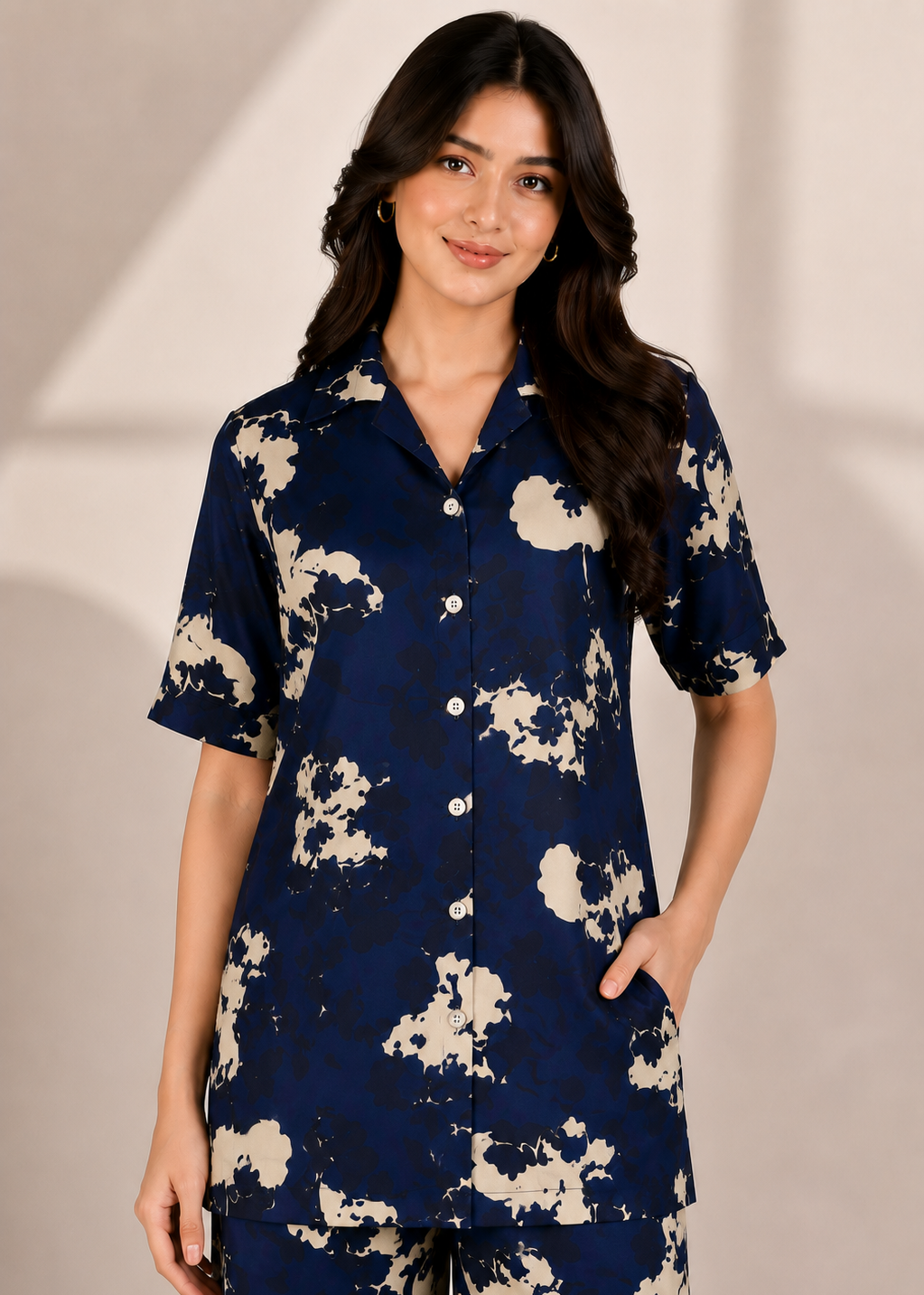 A woman modeling a navy blue short-sleeve button-up shirt and matching pants set with a beige abstract floral print, set against a neutral beige background.