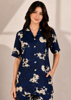 A woman modeling a navy blue short-sleeve button-up shirt and matching pants set with a beige abstract floral print, set against a neutral beige background.