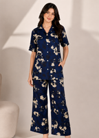 A woman modeling a matching two-piece pajama set with a navy blue base and beige floral print, featuring a short-sleeve button-up top and wide-leg pants.