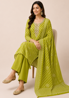 A woman seated on a chair wearing a bright yellow-green traditional outfit with matching pants and a draped scarf, photographed against a plain beige studio background.
