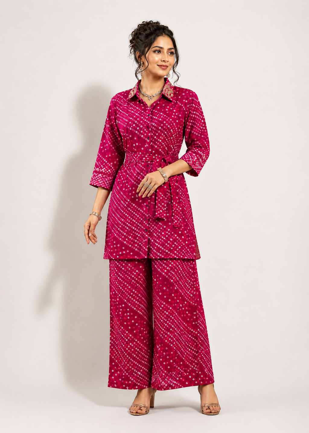 A woman modeling a bright pink two-piece kurta set with a white dotted pattern, featuring a collared wrap-style top with a tie belt and matching wide-leg pants, set against a plain light gray studio background.
