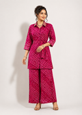 A woman modeling a bright pink two-piece kurta set with a white dotted pattern, featuring a collared wrap-style top with a tie belt and matching wide-leg pants, set against a plain light gray studio background.