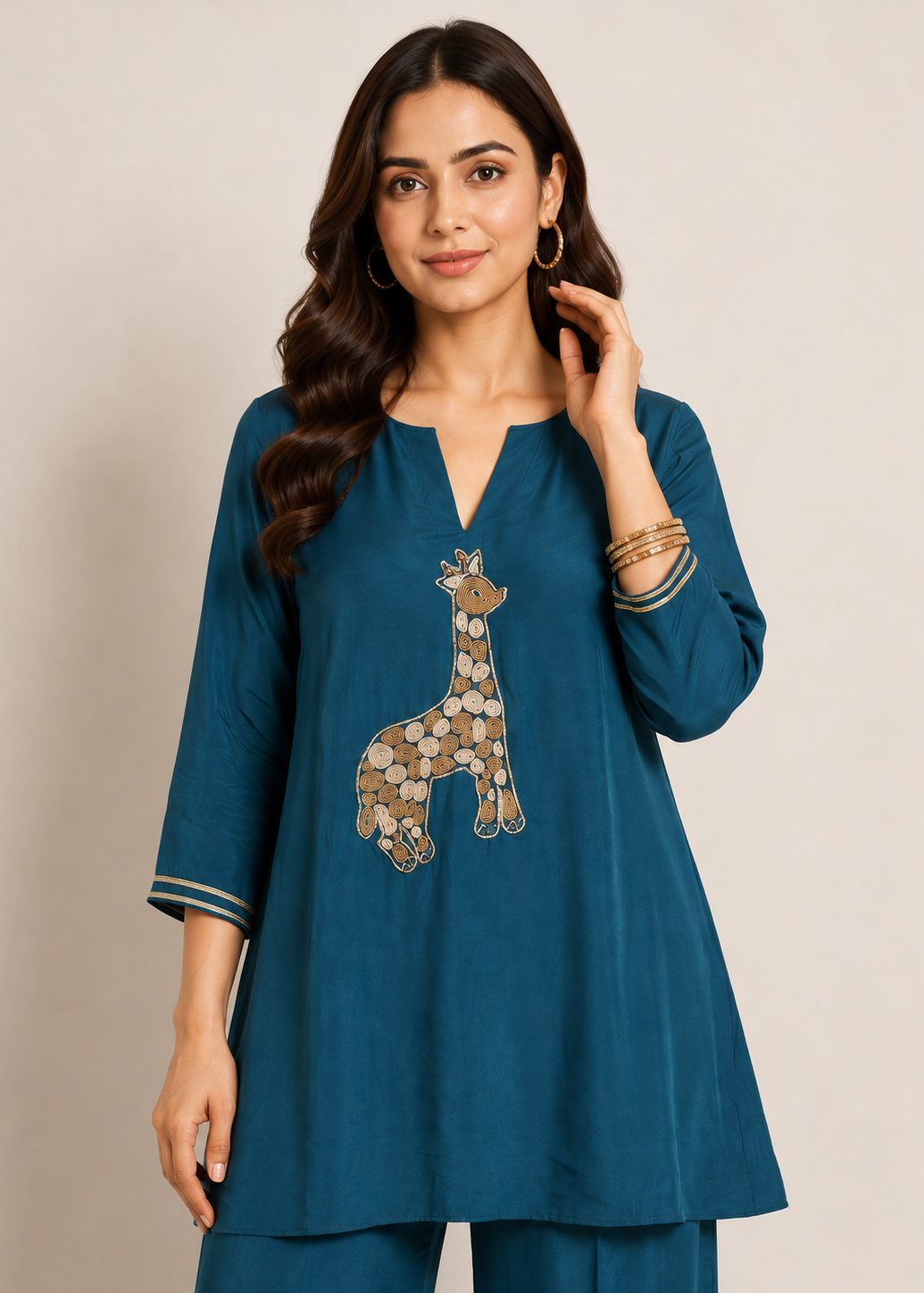 A woman modeling a teal blue kurta with a giraffe patchwork design on the front, paired with matching trousers, set against a plain beige studio background.