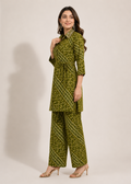 A woman modeling a matching olive green two-piece outfit with a small white floral print and white geometric border patterns, consisting of a long-sleeve tunic top with a tie belt and wide-leg pants, set against a plain beige studio background.