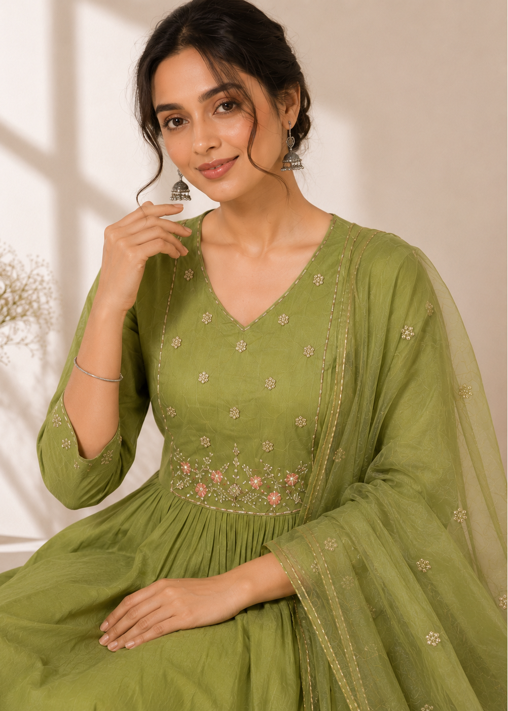 A woman wearing a sage green traditional Indian outfit with floral embroidery and a matching sheer dupatta, posed in a softly lit indoor setting.