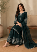 A woman models a dark teal green traditional South Asian outfit with white floral embroidery and silver mirror work, seated on a wooden bench in a warm, neutral-toned room.