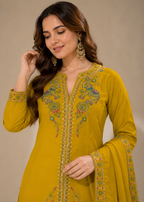 A woman wearing a mustard yellow traditional embroidered kurta with green, red, and gold detailing, paired with matching embroidered dupatta and gold earrings, set against a plain beige background.