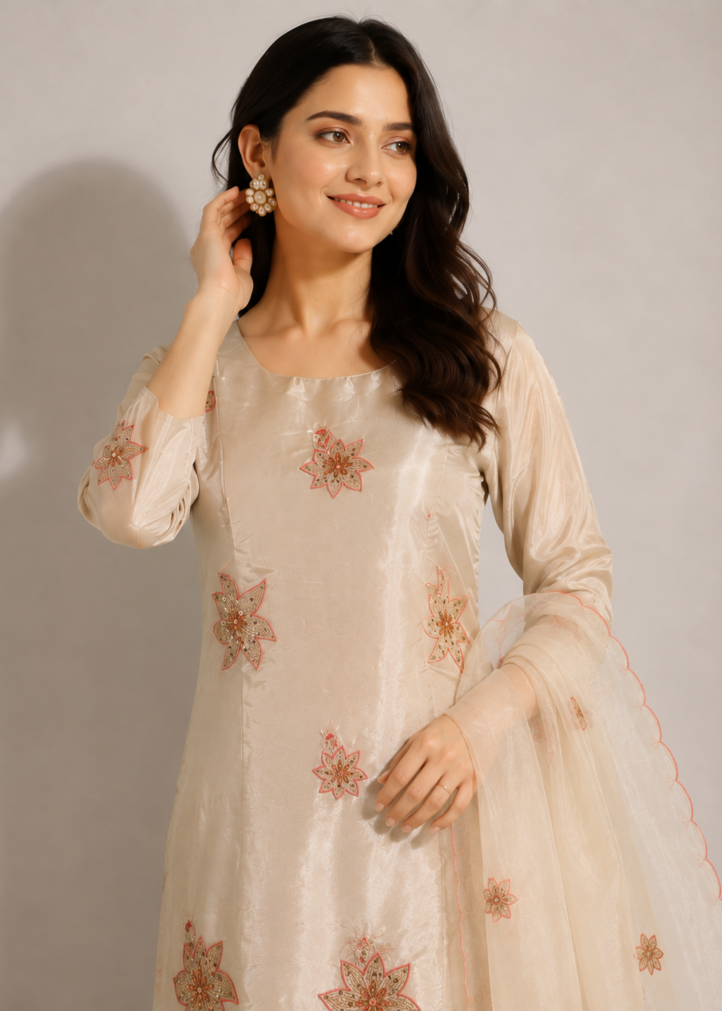 A woman wearing a light beige or cream-colored traditional kurta with pink floral embroidery and a matching sheer dupatta, posed against a neutral light gray studio background.