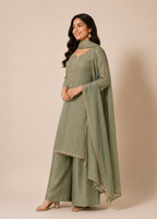 A woman modeling a sage green traditional Indian outfit with a matching dupatta, featuring subtle gold embroidery and a high-neck design, set against a solid beige studio background.