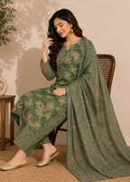 A woman seated on a wooden chair wearing an olive green floral salwar kameez suit with a matching dupatta, set against a warm beige wall with a small side table and plant.