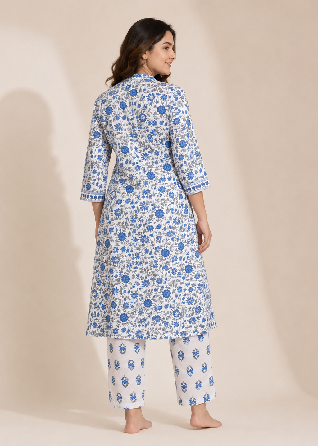 A woman modeling a white three-piece kurta set with blue floral block prints, shown from the back against a warm beige studio backdrop.