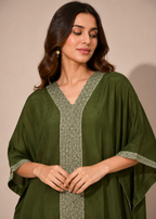 A woman wearing an olive green kaftan-style tunic with intricate silver and white embroidery along the V-neckline, front placket, and sleeve cuffs, posed against a solid beige studio background.