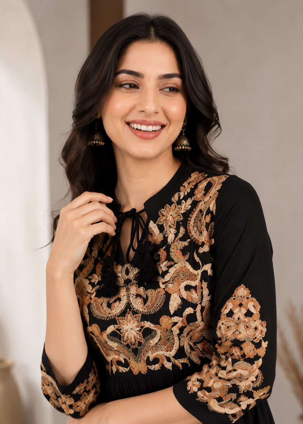 A woman wearing a black dress with ornate gold and beige floral embroidery on the bodice and sleeves, standing in a softly lit indoor setting.