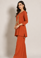 A woman modeling a rust-orange two-piece kurta set with a matching wide-leg pant. The kurta features a V-neck, three-quarter sleeves, and a decorative embroidered giraffe motif on the chest.