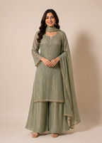 A woman modeling a sage green Indian ethnic suit set with a high-neck kurta, wide-leg pants, and a matching dupatta, set against a solid beige studio background.