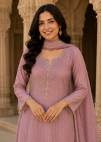 A woman wearing a dusty purple traditional Indian salwar kameez set with gold embroidery, standing in front of a stone archway.