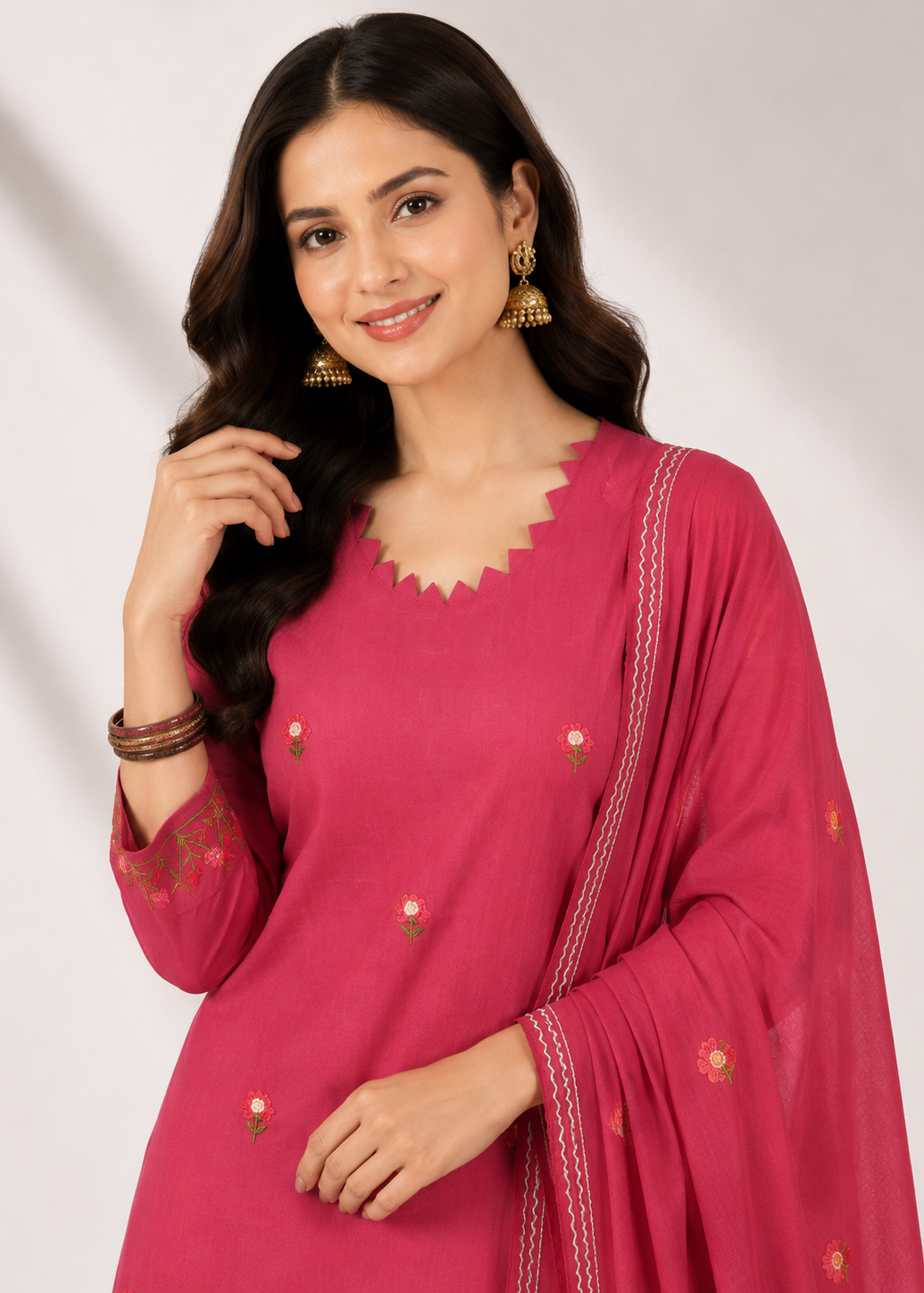 A woman modeling a bright pink traditional outfit with a scalloped V-neckline, embroidered details, and a matching sheer dupatta, set against a light gray studio background.