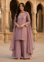 A woman models a lavender purple traditional Indian outfit with matching dupatta and wide-leg pants, standing in front of an ornate stone archway.