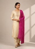 A woman modeling a traditional Indian outfit consisting of a cream-colored kurta with floral embroidery, matching trousers, and a bright magenta-pink patterned dupatta, set against a neutral beige studio background with soft window shadow projections.