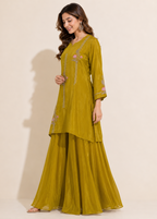 A woman modeling a three-piece olive green traditional Indian outfit with a tunic, flared pants, and embroidered details, set against a plain beige studio background.