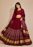 A woman modeling a deep maroon red Indian ethnic lehenga choli set with gold metallic borders and patterns, set against a plain beige studio background.
