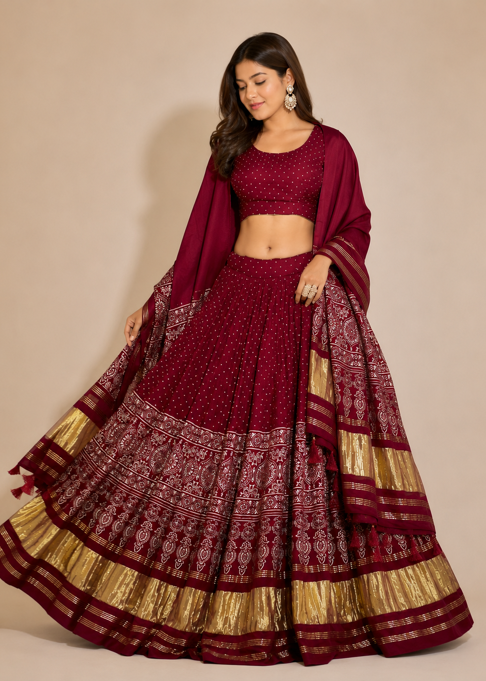A woman modeling a deep maroon red Indian ethnic lehenga choli set with gold metallic borders and patterns, set against a plain beige studio background.