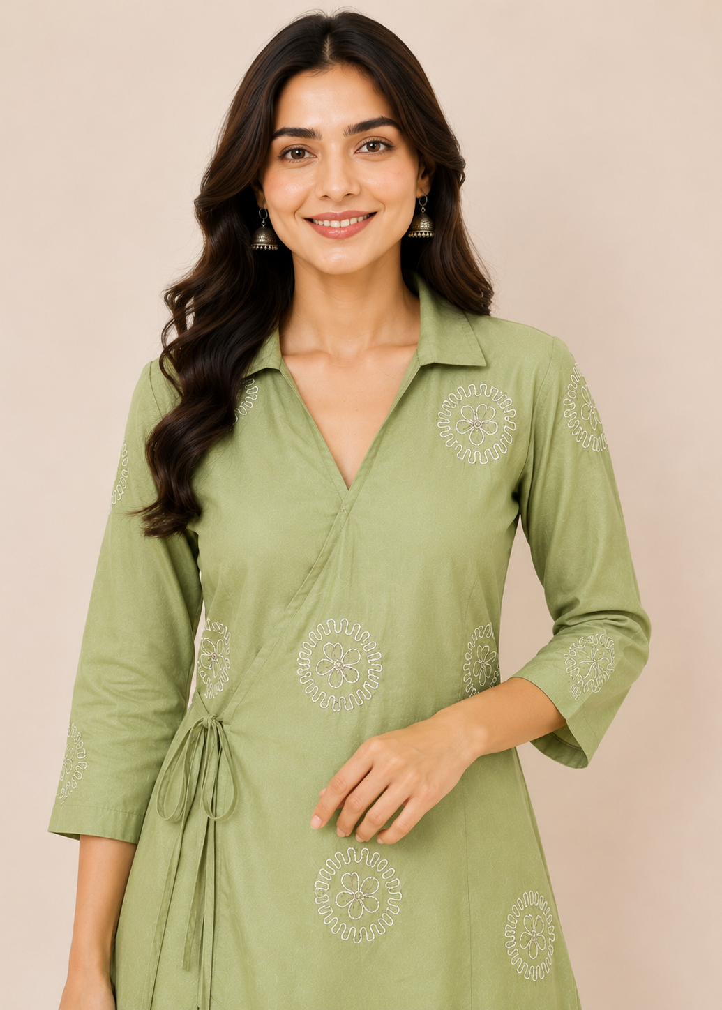 A woman wearing a sage green wrap-style kurta dress with white floral embroidery, standing against a solid beige background.