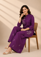 A woman sits on a wooden chair wearing a purple traditional outfit with white floral embroidery, photographed in a studio with warm beige walls and soft diagonal shadows.