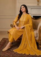 A woman seated on an ornate armchair in a traditional mustard-yellow three-piece outfit with matching dupatta, set in an elegant indoor room with a fireplace and patterned rug.