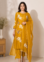 A woman modeling a mustard yellow three-piece traditional outfit with white floral embroidery, consisting of a long kurta, matching pants, and a draped dupatta, set against a warm beige studio background.