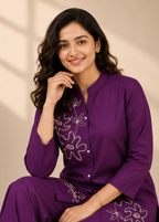 A woman wearing a purple traditional kurta set with white floral embroidery, posing against a warm beige background with soft shadow overlays.